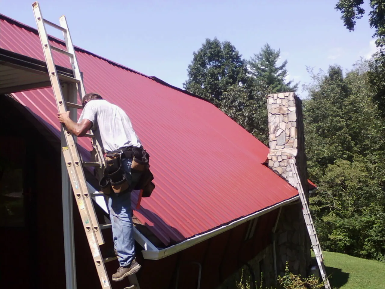 Roofer on a ladder beside a red metal roof and stone chimney — Soffit & Fascia Repair by Roofing Brothers in Northford
